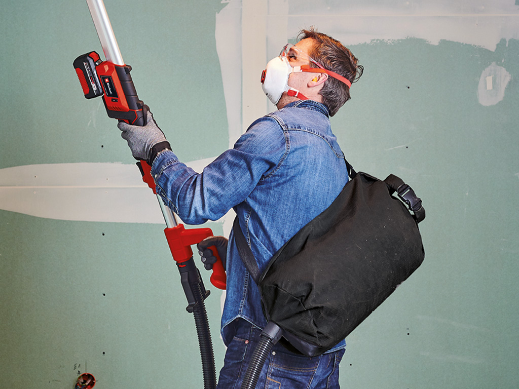 A man is sanding the wall and has a collecting bag on his back