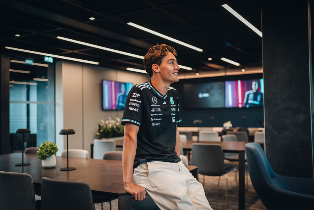 George Russell, Fahrer des Mercedes-AMG PETRONAS Formula One Teams, sitzt lässig auf einem Tisch in einem modernen, stilvollen Team-Hospitality- oder Bürobereich. Er lächelt und trägt das offizielle Teamshirt mit gut sichtbaren Sponsorenlogos wie Einhell, IWC und CrowdStrike. Auf Bildschirmen im Hintergrund ist sein Bild zu sehen.