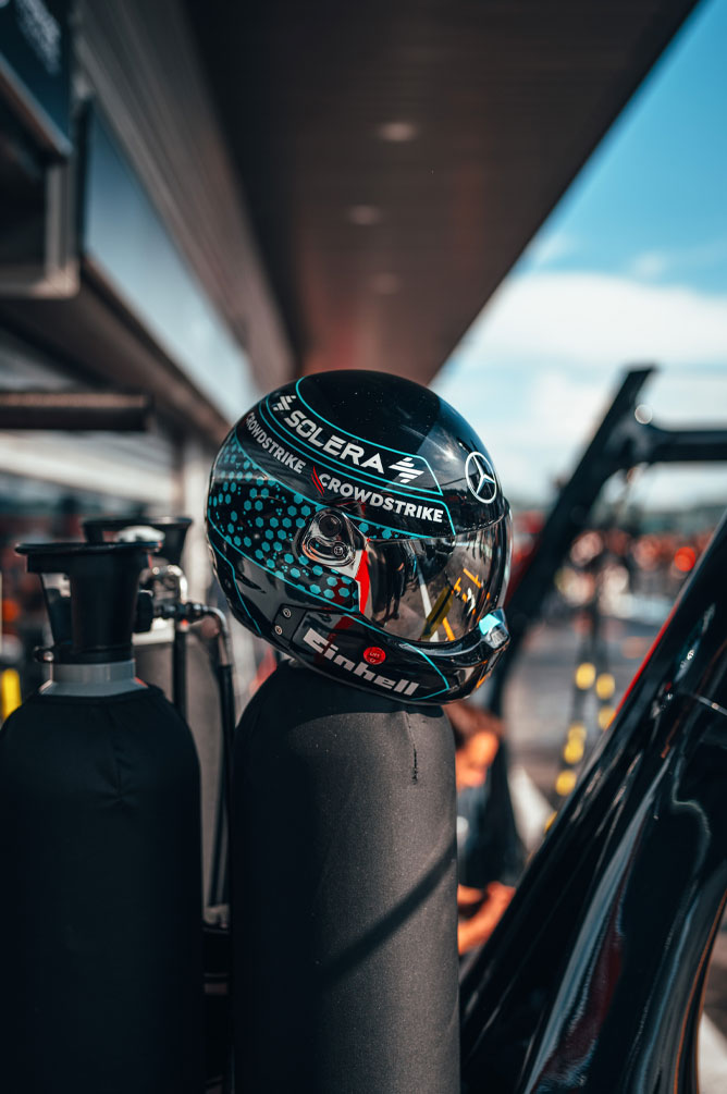 Ein Helm des Mercedes-AMG PETRONAS Formula One Teams mit gut sichtbaren Sponsorenlogos, darunter Einhell, CrowdStrike und Solera, ist auf einer Gasflasche in der Boxengasse platziert. Die Spiegelung im Visier zeigt die Umgebung der Garage.