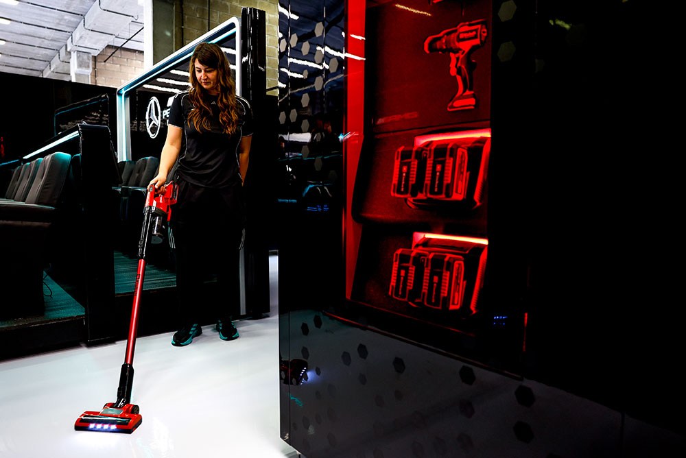 Eine Frau des Mercedes-AMG PETRONAS Formula One Teams reinigt einen glänzend weißen Boden mit einem kabellosen Staubsauger von Einhell. Rechts zeigen rot beleuchtete Regale mehrere Einhell Power X-Change Akkus und eine Akku-Bohrmaschine.