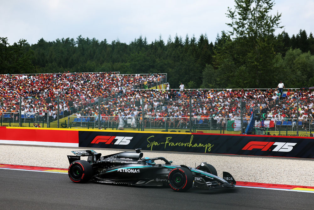 Ein Mercedes-AMG PETRONAS Formula One Wagen fährt auf dem Circuit de Spa-Francorchamps. Im Hintergrund sind volle Tribünen mit Fans zu sehen, die internationale Fahnen schwenken. Das Spa-Francorchamps-Logo ist auf der Barriere abgebildet.