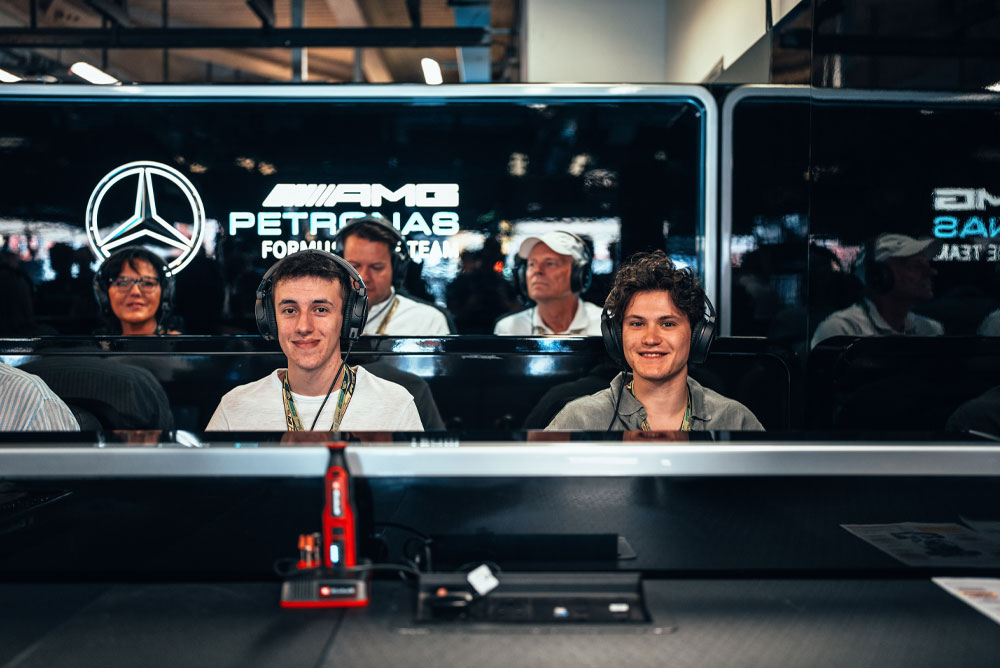 Zwei Männer sitzen in der Garage des Mercedes-AMG PETRONAS Formula One Teams, tragen Headsets und genießen das Behind-the-Scenes-Erlebnis. Im Vordergrund ist ein rotes Einhell-Gerät sichtbar.