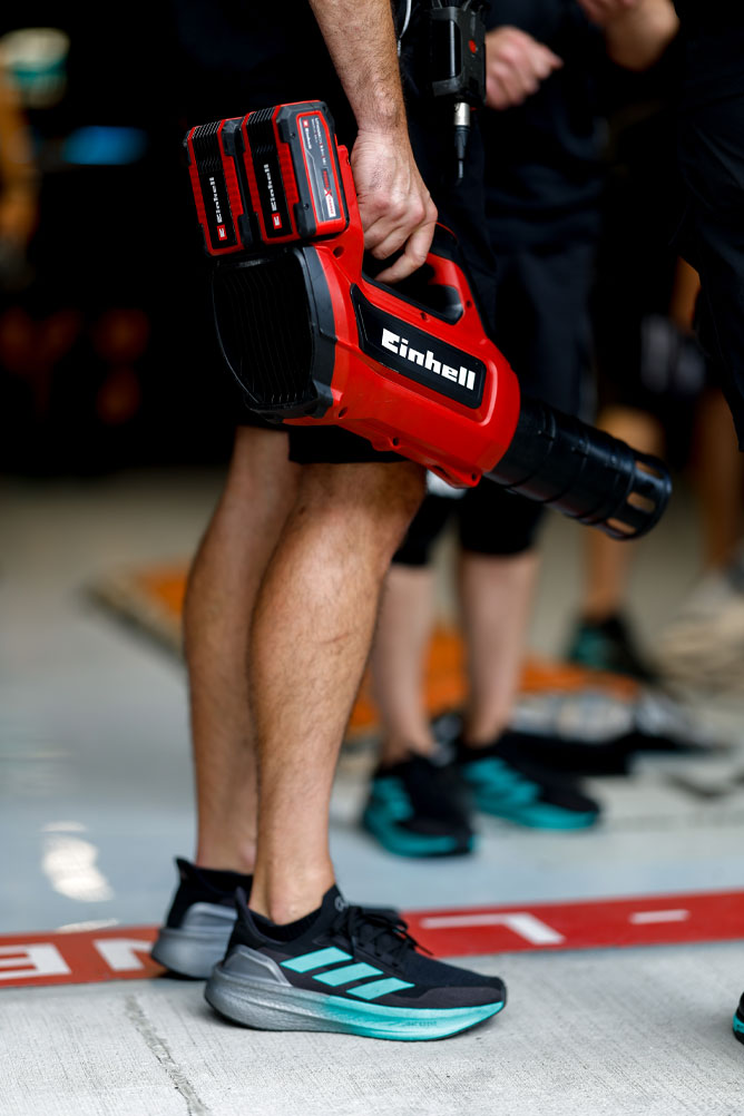 Ein Teammitglied hält ein rotes kabelloses Gebläse von Einhell mit zwei Power X-Change Akkus. Die Szene spielt sich im Boxenbereich während eines Formel-1-Events ab.