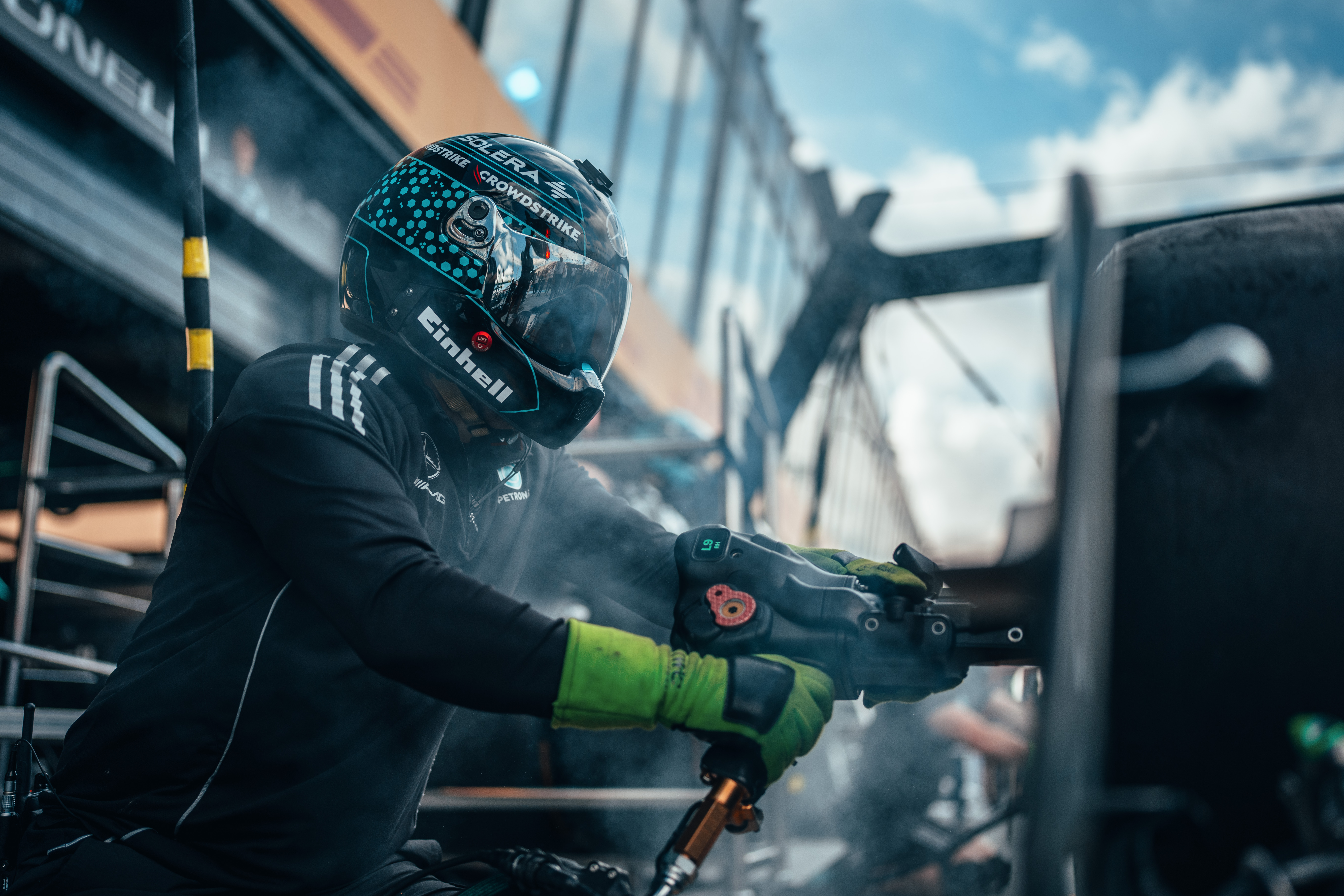 Ein Mitglied der Formel-1-Boxencrew trägt einen Helm und Handschuhe und benutzt während eines Boxenstopps eine pneumatische Radpistole, umgeben von Rauch und Ausrüstung.