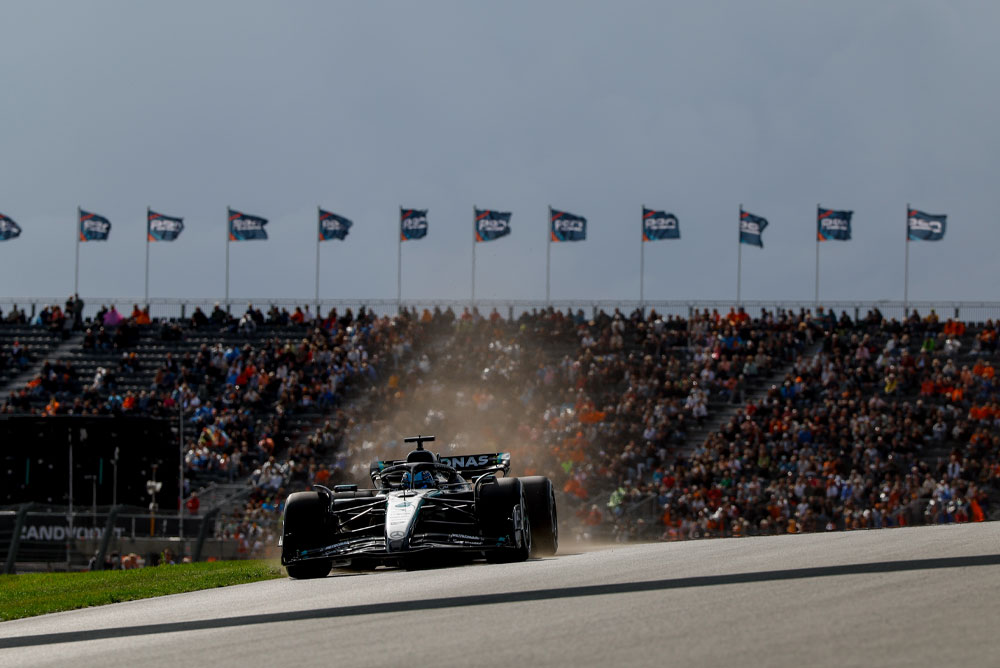 Ein Mercedes-AMG PETRONAS Formula One Wagen fährt während eines Rennens an vollen Tribünen vorbei. Fans schwenken Fahnen im Hintergrund und schaffen eine mitreißende Atmosphäre.
