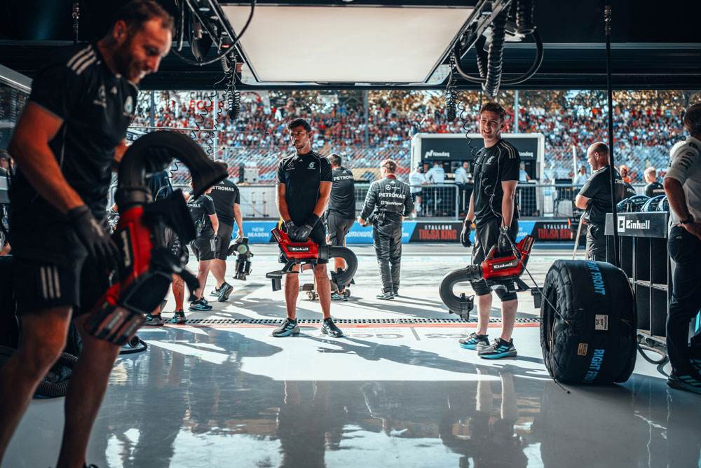 Mechaniker des Mercedes-AMG PETRONAS Formula One Teams bereiten sich in der Garage mit kabellosen Gebläsen von Einhell auf einen Boxenstopp vor. Im Hintergrund sind die mit Fans gefüllten Tribünen zu sehen.