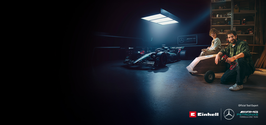 Ein Vater und Kind sind in einer Werkstatt mit einem selbstgebauten Seifenkistenauto. Im Hintergrund ein Mercedes-AMG PETRONAS Formel-1-Wagen und Einhell-Werkzeuge.