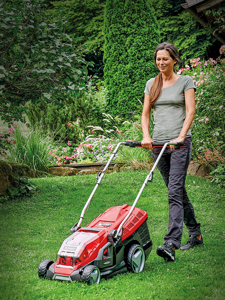 A woman is mowing the lawn in a green, blooming garden with a red Einhell lawn mower.