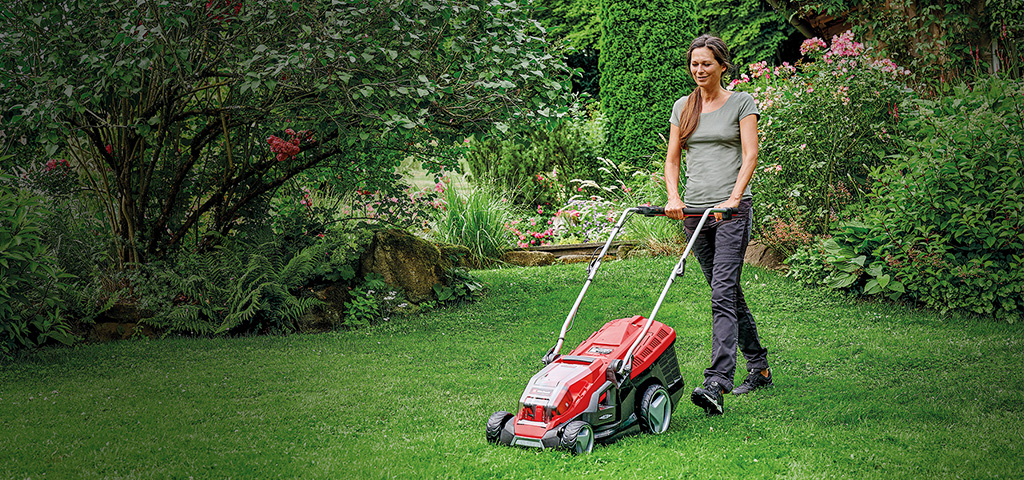 Žena sekačkou na trávu Einhell červené barvy kosí trávník v zelené, kvetoucí zahradě