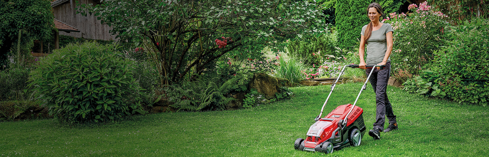 Žena sekačkou na trávu Einhell červené barvy kosí trávník v zelené, kvetoucí zahradě.
