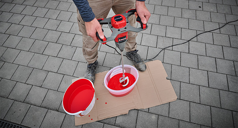Eine Person verwendet einen Rührer, um rote Farbe in einem Eimer auf einem Karton zu mischen.