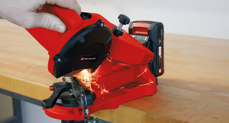 A person sharpens a saw chain using a red Einhell cordless chainsaw sharpener on a workbench.