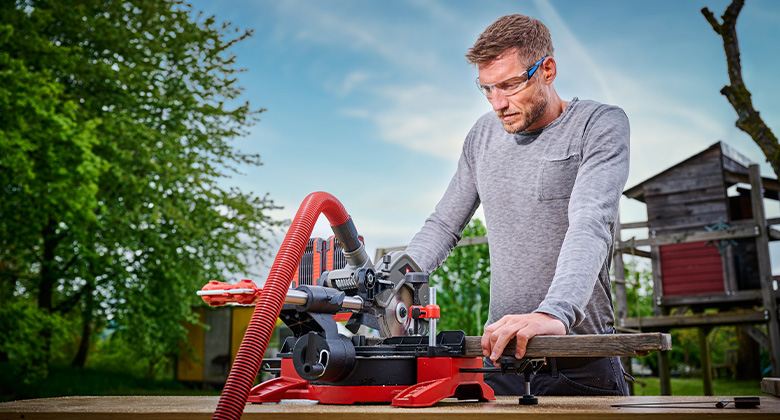 A man wearing safety goggles cuts wood outdoors using a red Einhell cordless mitre saw.