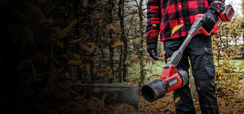 Eine Person trägt eine schwarze Hose, Handschuhe und eine rot-schwarz karierte Jacke und steht im Freien mit einem roten kabellosen Laubbläser von Einhell. Herbstlaub bedeckt den Boden, im Hintergrund sind Bäume zu sehen.