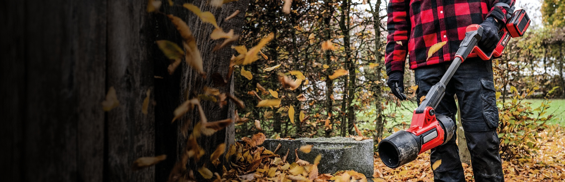 Eine Person trägt eine schwarze Hose, Handschuhe und eine rot-schwarz karierte Jacke und steht im Freien mit einem roten kabellosen Laubbläser von Einhell. Herbstlaub bedeckt den Boden, im Hintergrund sind Bäume zu sehen.