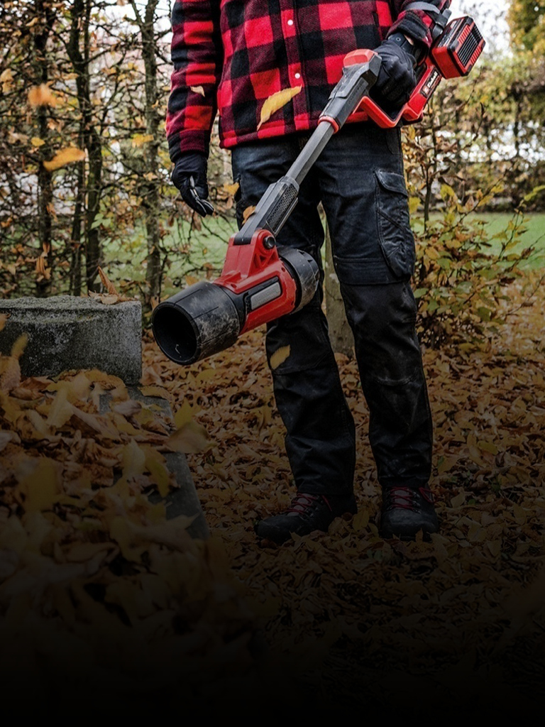 Eine Person trägt eine schwarze Hose, Handschuhe und eine rot-schwarz karierte Jacke und steht im Freien mit einem roten kabellosen Laubbläser von Einhell. Herbstlaub bedeckt den Boden, im Hintergrund sind Bäume zu sehen.